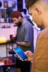 Caucasian young adult man holding smartphone with digital wallet app open, standing in tattoo studio while tattooed Caucasian young adult man working in background