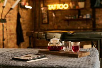 Tattoo studio interior showing glass teapot with red tea and two cups on wooden tray beside digital tablet and stylus on textured surface, tattoo bed and neon sign in background