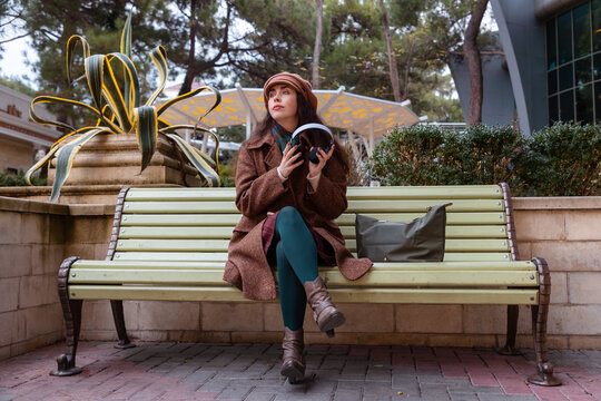 Wide shot of a Caucasian young woman in a hat and coat listening to music with headphones in a park, sitting on bench. Rest and relaxation alone