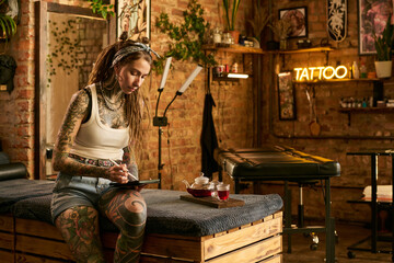 Caucasian young adult woman with extensive tattoos sitting on massage table drawing on digital tablet in tattoo studio, tea set on tray beside her, exposed brick walls in background