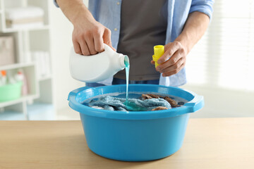 Man pouring detergent into plastic basin with towels at wooden table indoors, closeup