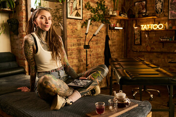 Portrait of young adult Caucasian woman with long hair and extensive tattoos sitting cross legged...