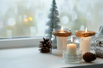 DIY Christmas lanterns. Burning candles in glass jars and cones on window sill indoors, closeup. Space for text