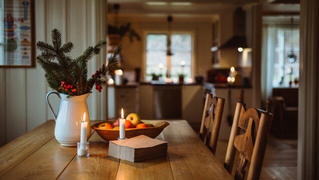 Cozy rustic dining table set for a holiday meal with candlelight, white jug of pine and red berries, bowl of apples and kraft napkins creating a warm festive interior ambiance
