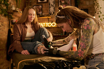 Woman with long hair sitting on tattoo table receiving leg tattoo from tattooed Caucasian young adult woman artist concentrating on work in studio