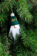 Handmade gnome ornament with green knit hat and white beard on fir branches with authentic natural look, festive mood and soft copy space. Concept of cozy holiday decoration, winter magic.