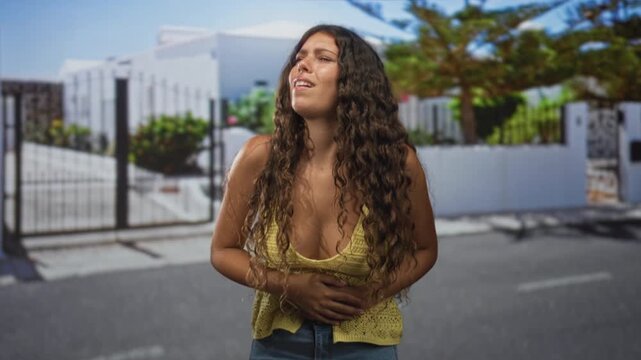 Woman clutching bare stomach and wincing with hands on belly while standing by a gated building on street; pain nausea.