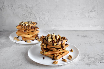 Golden waffles stacked on white plates, topped with banana slices, walnuts, and chocolate drizzle, creating a delicious and visually appealing breakfast or dessert scene.