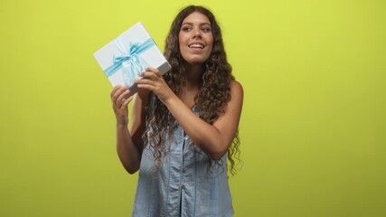 Woman holds a white gift box with blue ribbon and shakes it while listening in a studio; anticipation discovery.