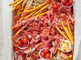 A colorful platter features a variety of cured meats, cheeses, cherry tomatoes, nuts, and breadsticks. Perfect for gatherings, it displays an inviting assortment of flavors and textures