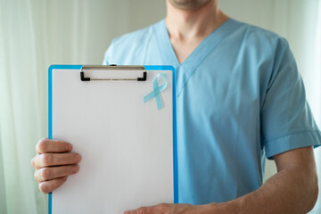 Male healthcare professional in uniform with blue ribbon symbol of prostate cancer awareness holding clipboard with empty paper for notes and reminders in November.