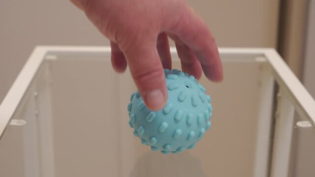 A small, light blue textured rubber or silicone ball is released from a hand and falls onto a clear glass tabletop. The soft, bouncy material makes a very quiet thump as it contacts the hard surface.