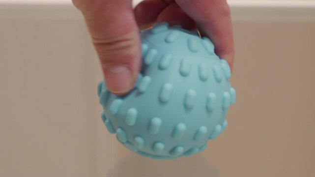 A textured rubber or silicone ball is squeezed firmly by a hand. The pressure forces air out of the toy, resulting in a distinct, high-pitched squeaking sound.