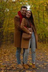 Happy couple hugging and smiling in autumn park, wearing matching brown coats and red accents, standing on a path covered with yellow leaves surrounded by fall trees.
