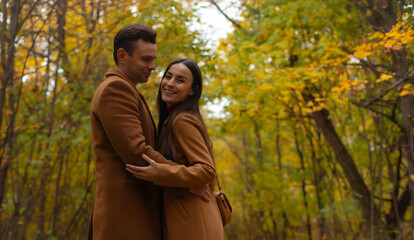 Happy couple embracing in autumn forest, both wearing brown coats, smiling and enjoying romantic moment surrounded by colorful fall foliage and natural beauty.