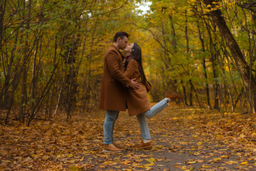 Romantic couple kissing in autumn forest, both wearing brown coats and jeans, surrounded by colorful fall leaves, expressing love and happiness in a scenic park.