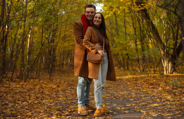 Smiling couple standing and hugging in autumn park, dressed in matching brown coats and jeans, surrounded by colorful yellow leaves and warm fall atmosphere.