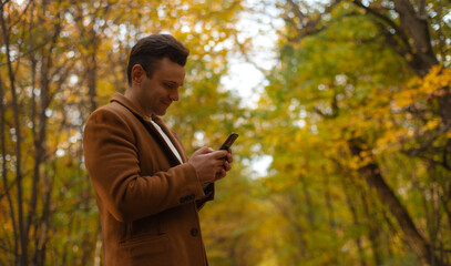 Smiling man in brown coat using smartphone in autumn forest, standing among golden leaves and trees, enjoying online communication and nature during fall season.