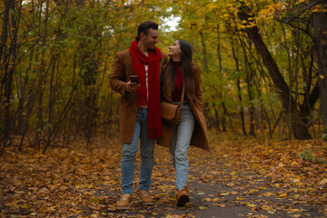 Happy couple walking together in autumn park, smiling and looking at each other, dressed in brown coats and jeans, enjoying romantic fall day surrounded by colorful leaves.