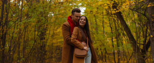 Smiling couple embracing in autumn forest, dressed in brown coats and red accents, enjoying romantic walk surrounded by colorful fall foliage and golden leaves.