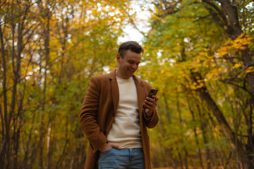 Smiling man in brown coat using smartphone in autumn park, enjoying communication and nature, standing among colorful trees and fallen leaves during warm fall day.