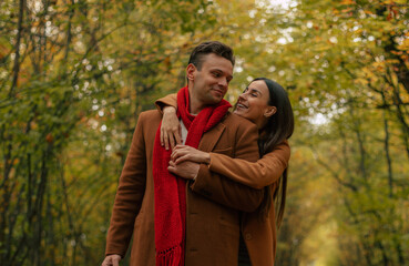 Smiling couple in brown coats embracing in autumn park, woman hugging man from behind, both enjoying warm fall day surrounded by golden leaves and romantic atmosphere.