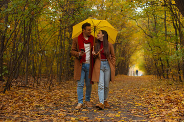 Happy couple walking under yellow umbrella in autumn park, dressed in matching brown coats and red accents, smiling and enjoying romantic walk surrounded by colorful fall foliage.