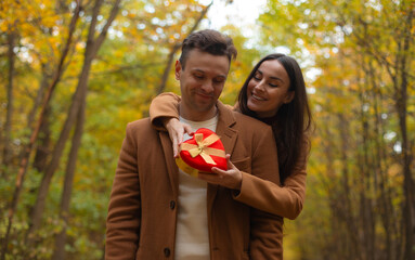 Romantic couple celebrating Valentine’s Day in an autumn forest. Woman surprises her partner with a heart-shaped gift box as they share a tender, happy moment together.