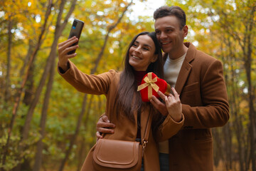 Happy couple taking a selfie on Valentine’s Day in an autumn forest. Woman holds a heart-shaped gift box while they smile and enjoy a romantic moment together.