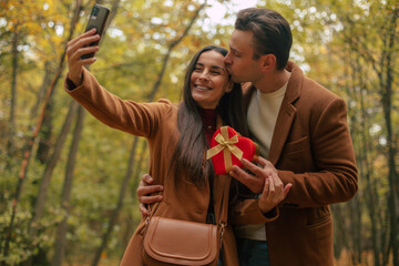 Happy couple celebrating Valentine’s Day in an autumn forest, taking a selfie while the man kisses the woman. She holds a heart-shaped gift box, capturing a romantic moment together.