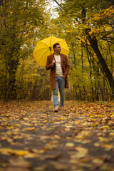 Young man walking in autumn park with yellow umbrella, wearing brown coat and jeans, enjoying peaceful fall weather surrounded by colorful foliage and trees.