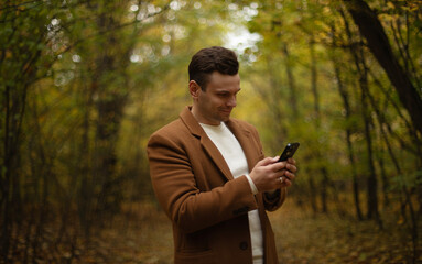 Smiling young man in brown coat using smartphone in autumn forest, standing among colorful trees, enjoying nature and technology during outdoor walk.