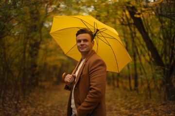 Smiling young man in brown coat holding yellow umbrella in autumn forest, looking at camera, enjoying peaceful fall day surrounded by colorful foliage and trees.