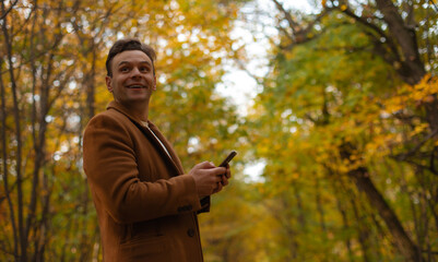 Smiling man in brown coat using smartphone in autumn forest, looking up with joy and optimism while surrounded by colorful fall foliage and warm natural light.