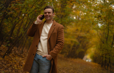 Smiling man in brown coat talking on smartphone while walking through autumn forest, enjoying conversation surrounded by yellow fall foliage and natural atmosphere.