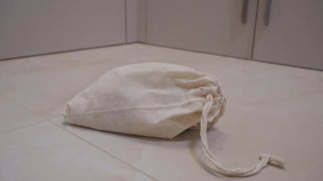 A cotton drawstring bag containing hard wooden blocks or similar objects is dropped onto a tiled floor or surface. The impact creates a distinctive muffled thud and a rattle.