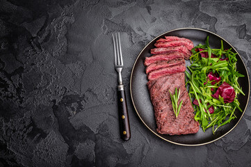 Grilled and sliced Flank beef steak on plate with salad. black background. top view