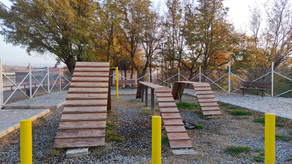 Wooden dog agility ramps and obstacles in an outdoor training area, surrounded by autumn trees and fenced pathways.