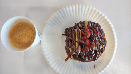 Top down view of a pancake topped with strawberries, banana slices, and chocolate drizzle, served on a paper plate beside a cup of frothy coffee.