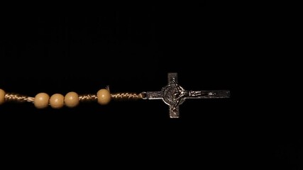 A metal cross on a rosary hangs in the darkness against a black background. A vertical, endlessly looping video.