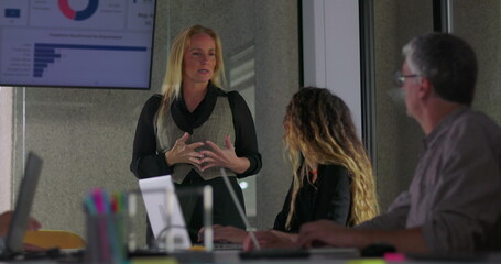Blonde businesswoman giving presentation to team while standing in office meeting room with data charts visible on screen and colleagues listening attentively
