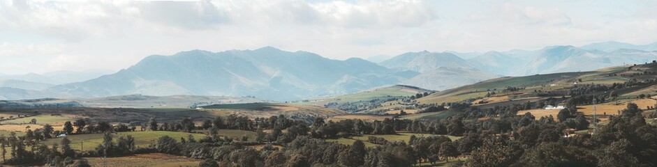 Obraz premium Wide telephoto panorama of rolling green hills, patchwork farmland and distant rugged mountains in soft light along Camino de Santiago in Asturias, capturing serene rural landscape beauty