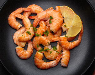 Cooked shrimps prawns arranged on a black plate with lemon wedges and chopped parsley, elegant top view seafood appetizer on dark background