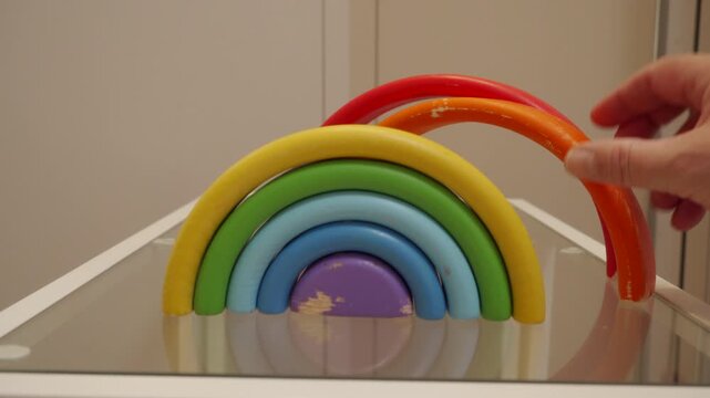 A wooden rainbow stacking toy is being completed as a hand places the final painted arch on top. The wood arcs make a quick, soft clack or tap as they nestle against the polished glass shelf.