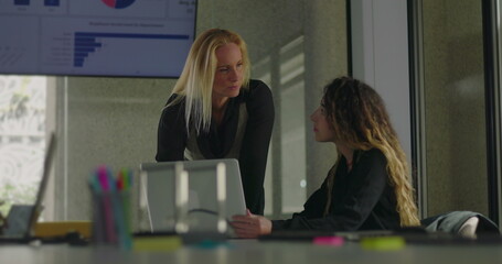 Blonde businesswoman leaning over desk to explain point to seated colleague during close collaboration in office with data chart on display