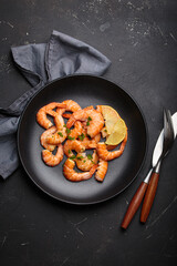Cooked shrimps prawns arranged on a black plate with lemon wedges and chopped parsley, elegant top view seafood appetizer on dark background