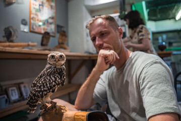 A speckled owl perches on a gloved stand as a man watches in a modern Hiroshima owl cafe. Indoor perch displays and multiple owls set a warm, educational tone.