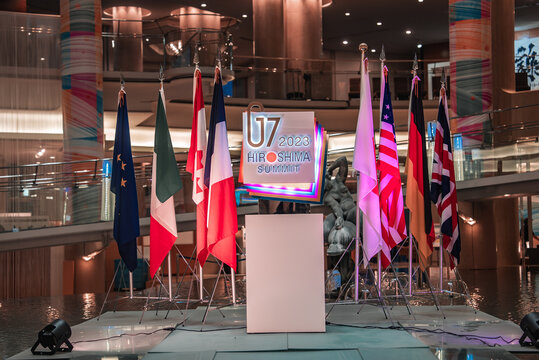 A podium stands in a modern venue in Hiroshima, Japan, for U7 2023. Flags of the EU, Italy, Canada, France, Japan, the United States, and the United Kingdom frame the stage.