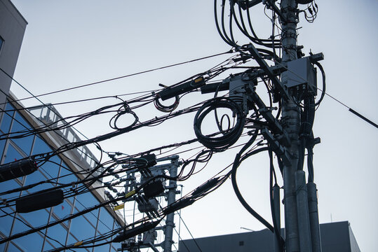 Overhead cables wrap a concrete power pole with loops, boxes, and transformers in Hiroshima, Japan, as a glass facade reflects the web in bright daytime light. - Powered by Adobe