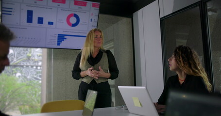Blonde businesswoman pointing at chart on large screen while explaining data to coworkers in formal office setting with laptops on table